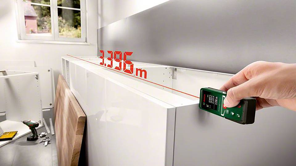 Eine Person misst mit einem Laser-Entfernungsmesser die Länge eines Küchenschranks, Anzeige zeigt '1,396 m'. Im Hintergrund ein Fenster.