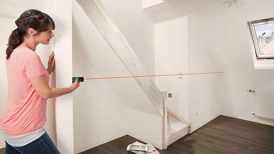 Eine Frau misst mit einem Laser-Entfernungsmesser die Breite eines Raums mit Dachschräge. Der Laserstrahl ist sichtbar an der Wand.