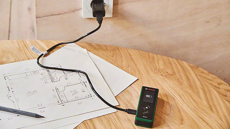 Ein digitaler Entfernungsmesser lädt an einer Steckdose auf einem Holztisch. Daneben liegt ein Bauplan mit Bleistift.