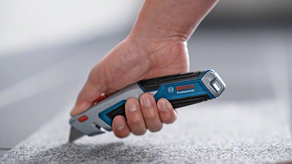 Eine Hand hält ein Bosch Professional Cuttermesser, das über einen grauen Teppichboden geführt wird.
