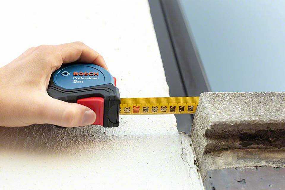 Eine Hand hält ein 5-Meter-Maßband der Marke Bosch und misst die Breite eines Fensterrahmens.