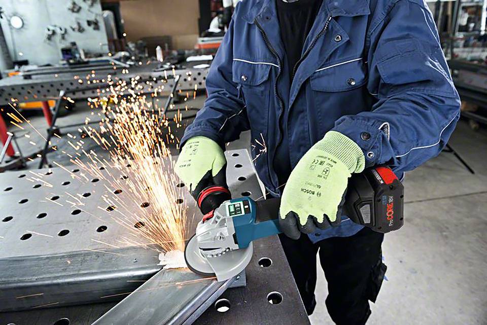 Person schleift Metallstück mit einer elektrischen Winkelschleifmaschine, Funken sprühen. Sie trägt blaue Jacke und grüne Handschuhe.