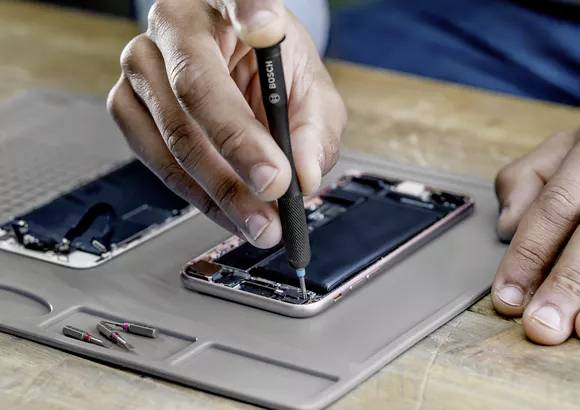 Person repariert ein Smartphone mit einem Schraubendreher; Ersatzteile und Werkzeuge liegen daneben auf einer Matte.