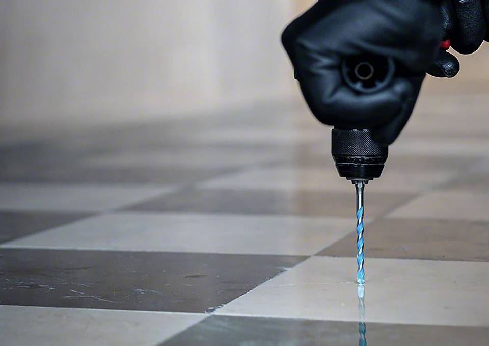 Eine Person bohrt mit einer Bohrmaschine ein Loch in eine schwarz-weiß karierte Bodenfliese, wobei sie schwarze Handschuhe trägt.