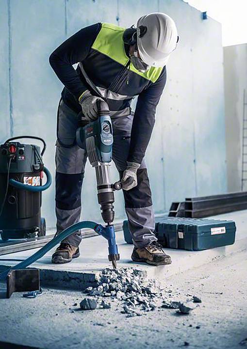 Arbeiter mit Schutzhelm benutzt einen Presslufthammer, um Beton zu durchbohren. Staubsauger und Werkzeugkasten im Hintergrund.