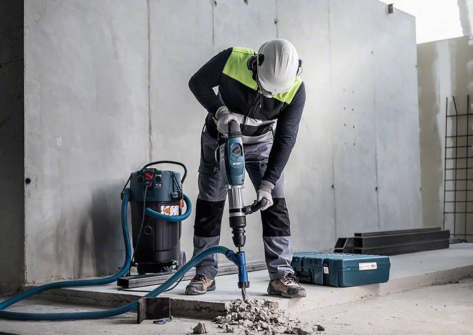 Arbeiter mit Schutzkleidung nutzt einen Bohrhammer, um Betonboden zu durchbrechen. Im Hintergrund steht ein Industriedammsauger.
