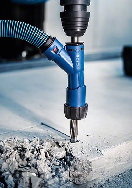 Ein blauer Bohrer mit Staubabsaugung bohrt in einen Betonboden. Staub und Betonstücke um den Bohrer verteilt, in einer Werkstattumgebung.