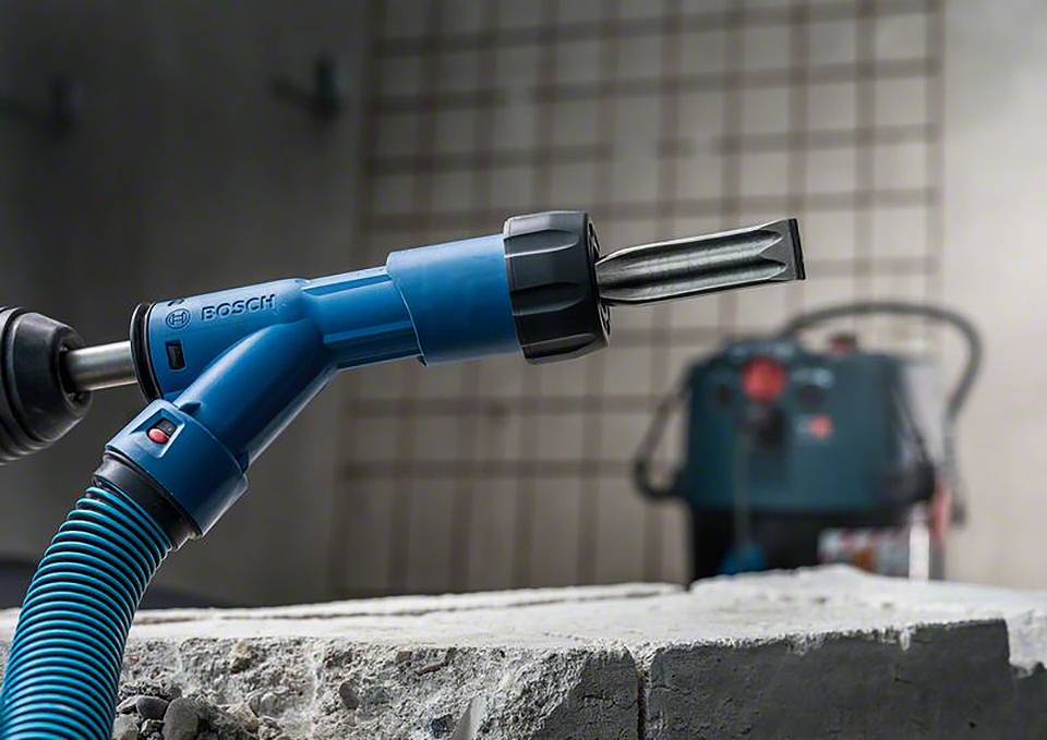 Ein blauer Bohrhammer bearbeitet Beton in einer Werkstatt. Im Hintergrund ist unscharf ein weiterer Bohrer oder Werkzeug erkennbar.