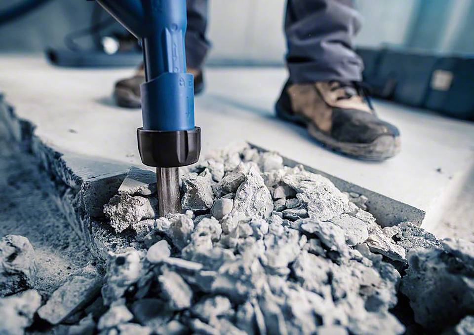 Eine Person verwendet einen elektrischen Presslufthammer, um Beton zu zerbrechen. Nahaufnahme von Schuhen und Werkzeug in Aktion.