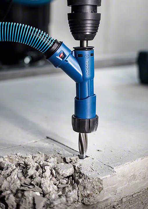 Ein blauer Bohrer bohrt in Beton, umgeben von Schutt. Ein Vakuumschlauch ist an den Bohrer angeschlossen, um Staub zu entfernen.