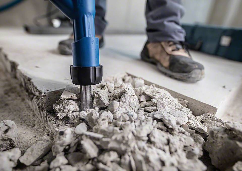 Eine Person arbeitet mit einem blauen Abbruchhammer und zerschlägt Betonboden. Staub und Trümmerstücke liegen herum.