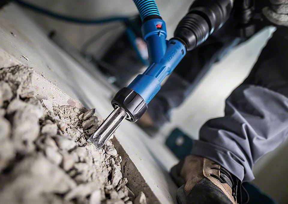 Eine Person verwendet einen blauen Bohrhammer, um Beton zu zerkleinern. Schutzkleidung und feste Schuhe sind sichtbar.