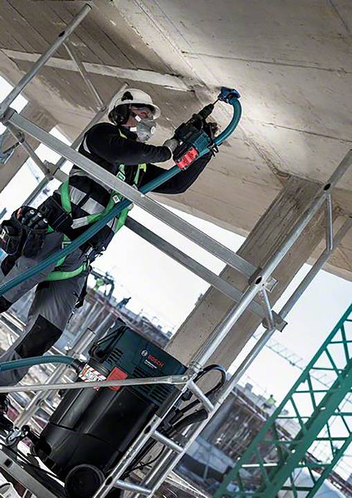 Bauarbeiter in Sicherheitskleidung steht auf einem Gerüst und bohrt in eine Betondecke, Baugeräte im Hintergrund.