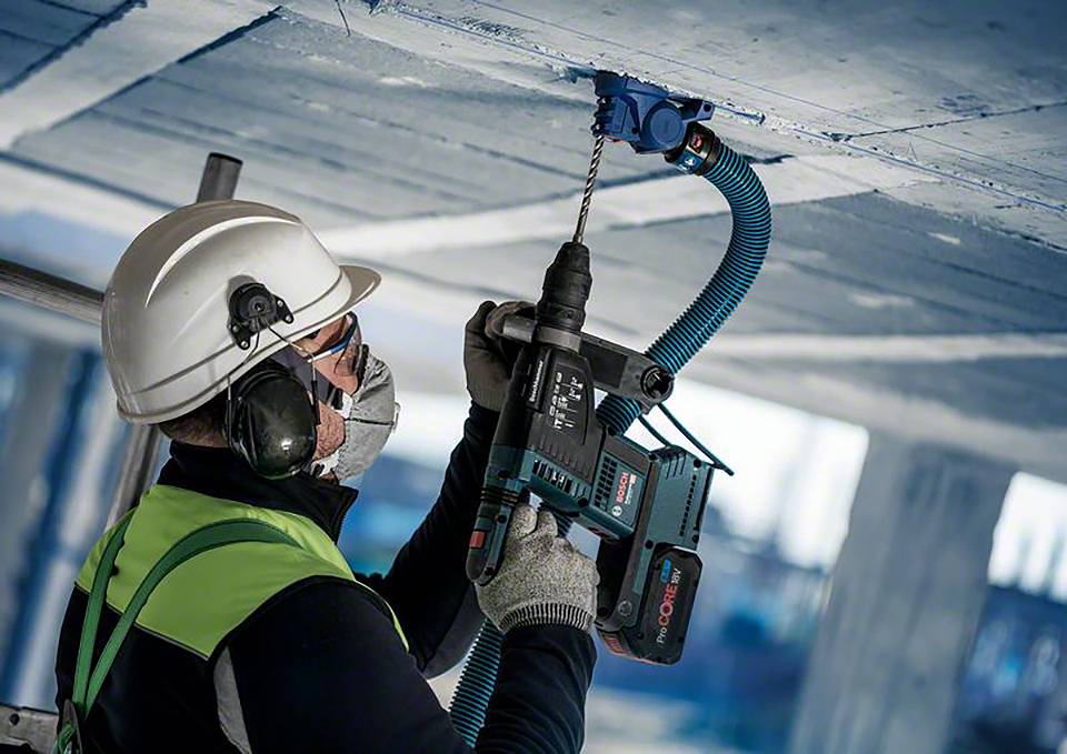 Ein Bauarbeiter mit Schutzhelm und Weste bohrt mit einer elektrischen Bohrmaschine in eine Betonoberfläche über Kopf.