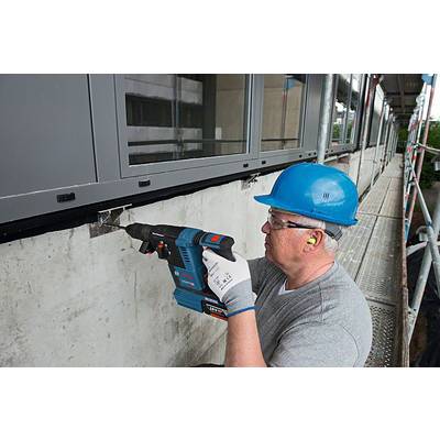 Ein Arbeiter in Sicherheitsausrüstung benutzt eine Bohrmaschine, um eine Befestigung an einem Betonfundament unter Fenstern anzubringen.