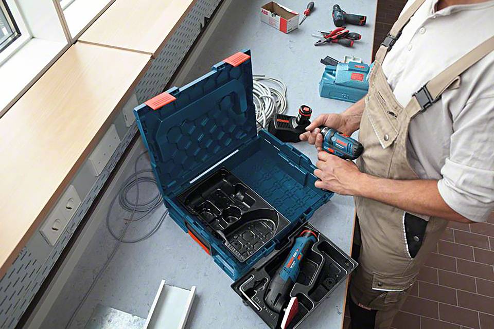 Ein Handwerker steht an einem Tisch, hält einen Akkuschrauber und öffnet eine Werkzeugkiste. Weitere Werkzeuge liegen bereit.