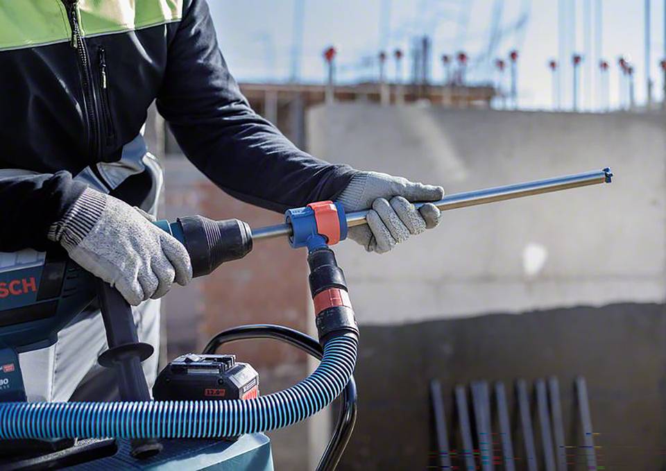 Arbeiter verwendet einen Elektroschrauber mit Staubsaugaufsatz auf einer Baustelle. Hintergrund zeigt Betonstruktur und Metallstangen.