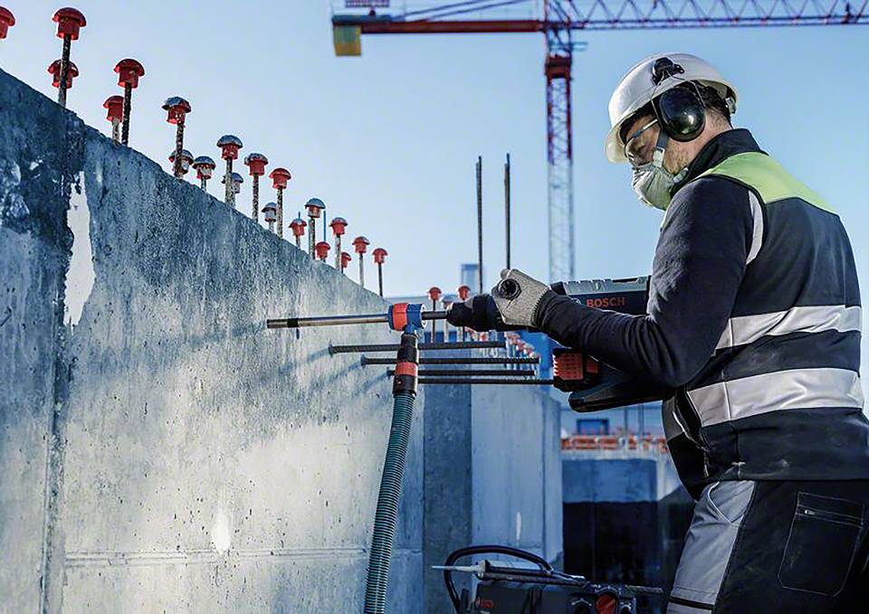 Ein Arbeiter in Schutzausrüstung bohrt mit einem elektrischen Bohrhammer in eine Betonwand auf einer Baustelle, Kräne im Hintergrund.