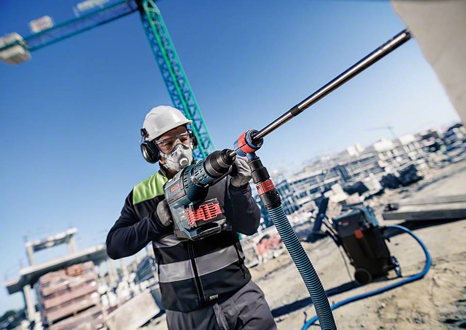 Ein Bauarbeiter mit Schutzkleidung und -maske nutzt einen Bohrhammer auf einer Baustelle, im Hintergrund sind ein Kran und Gebäude.