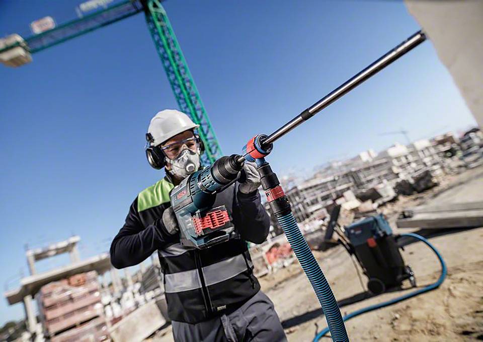 Ein Bauarbeiter mit Schutzhelm und Maske benutzt eine Bohrmaschine auf einer Baustelle, im Hintergrund sind Kräne und Gebäudestrukturen sichtbar.