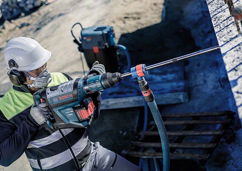 Eine Person arbeitet draußen mit einer Bosch-Bohrmaschine an einer Betonwand, trägt Schutzkleidung, Helm, Brille und Atemschutz.