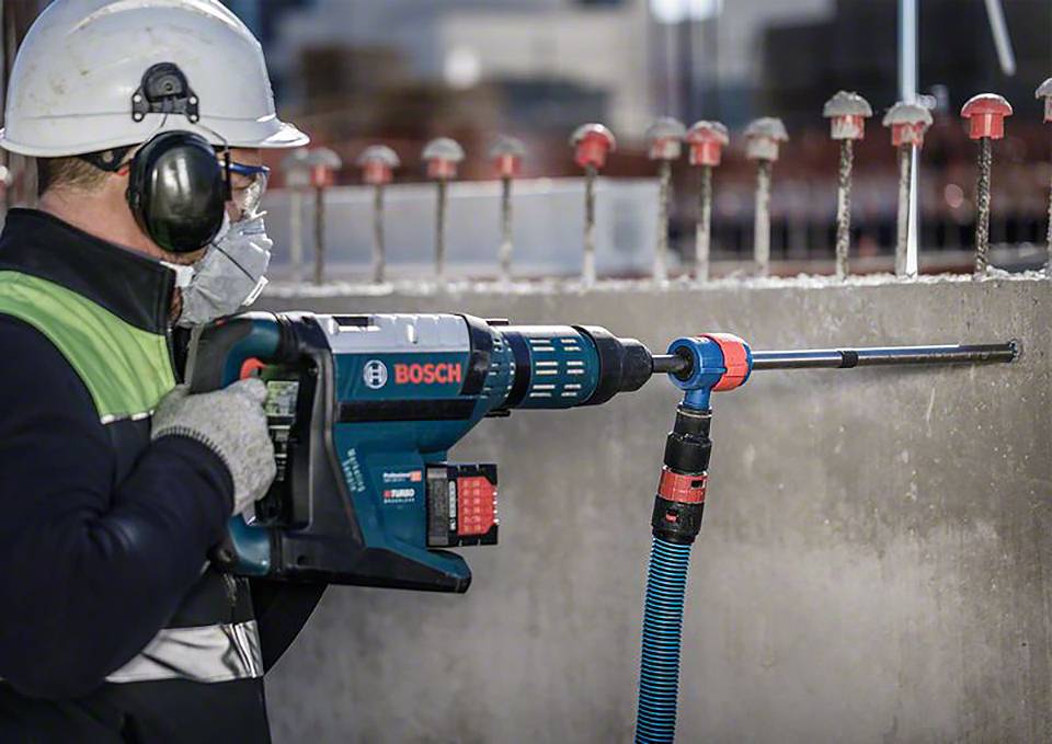 Eine Person in Arbeitskleidung und Schutzhelm bohrt mit einer Bosch-Bohrmaschine ein Loch in eine Betonwand auf einer Baustelle.