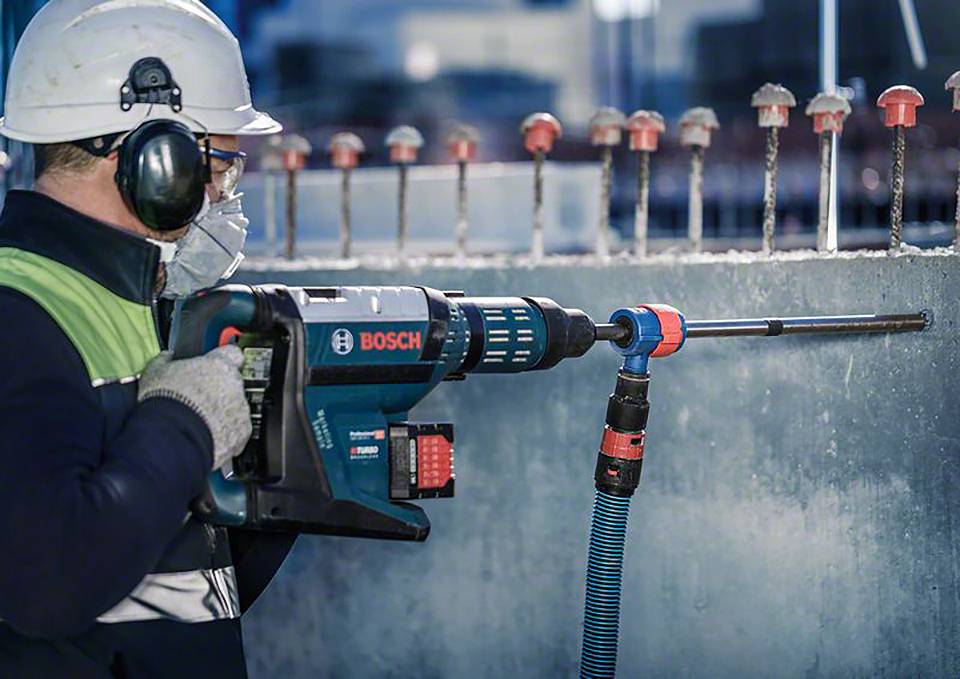 Ein Arbeiter trägt Helm und Gehörschutz, während er mit einer Bohrmaschine Löcher in eine Betonwand bohrt.