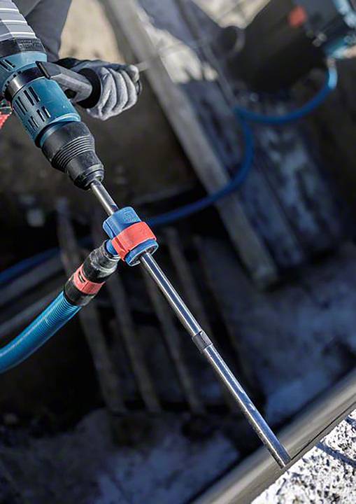 Eine Person bedient einen blauen Bohrhammer mit einem langen Bohrer. Im Hintergrund sind Baustellenmaterialien erkennbar.