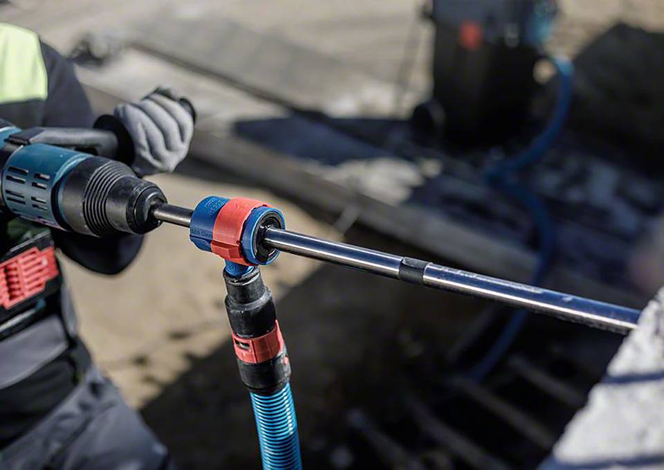 Ein Arbeiter bohrt mit einer elektrischen Bohrmaschine in eine Betonoberfläche. Schutzkleidung und Handschuhe sind zu sehen.