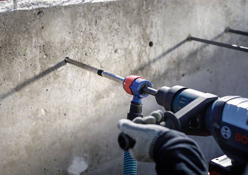Person verwendet einen Bohrhammer mit blauem Gehäuse, um ein Loch in eine Betonwand zu bohren. Eine blaue Spiralschlauchleitung ist angeschlossen.