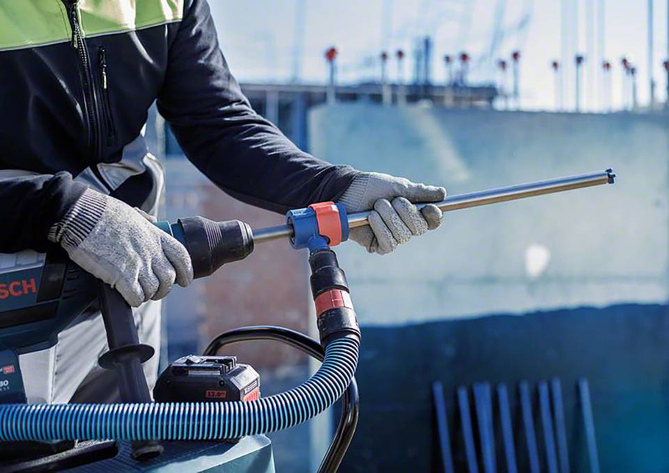 Arbeiter mit Schutzkleidung hält ein Elektrowerkzeug auf einer Baustelle. Im Hintergrund sind unscharf Baugerüste zu sehen.