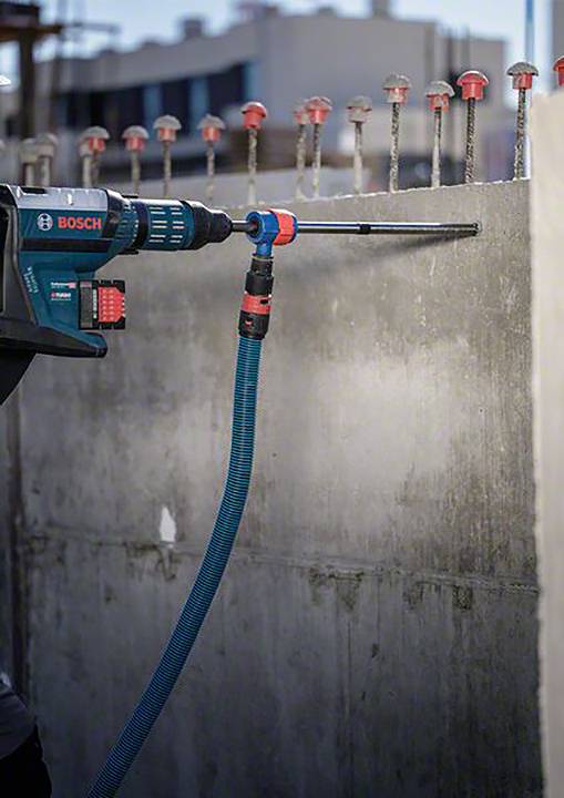 Eine elektrische Bohrmaschine bohrt ein Loch in eine Betonwand, dabei werden Stahlstangen sichtbar. Ein Staubsauger ist an das Werkzeug angeschlossen.