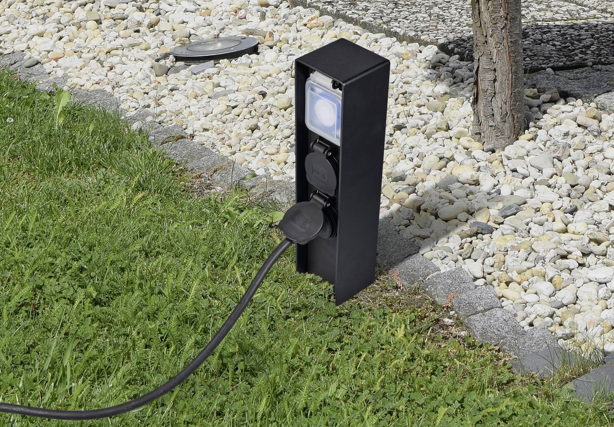Schwarze Außensteckdose im Garten, an einen Baum montiert, mit eingestecktem Kabel. Umgebung aus Gras und Kieselsteinen.