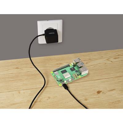 'Raspberry Pi' Computerplatine auf einem Holztisch, verbunden mit einem Stromadapter in einer Wandsteckdose.
