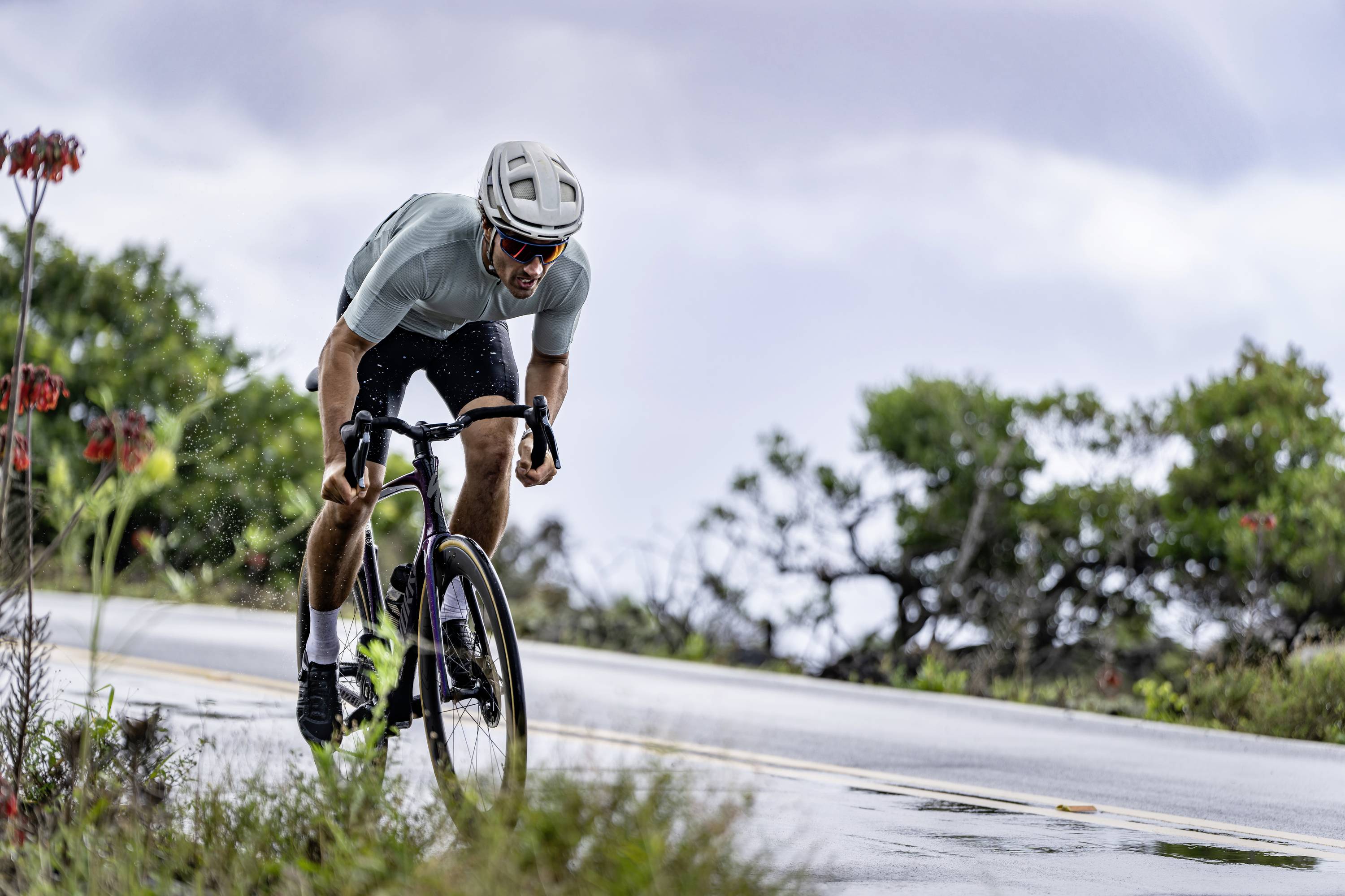 Ein Radfahrer in sportlicher Kleidung fährt bei Regen eine Straße entlang, umgeben von grüner Vegetation und bewölktem Himmel.