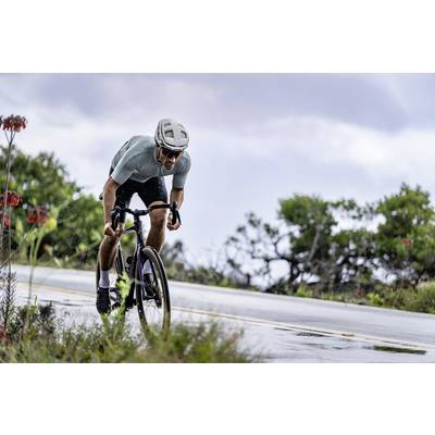 Ein Radfahrer in sportlicher Kleidung fährt bei Regen eine Straße entlang, umgeben von grüner Vegetation und bewölktem Himmel.