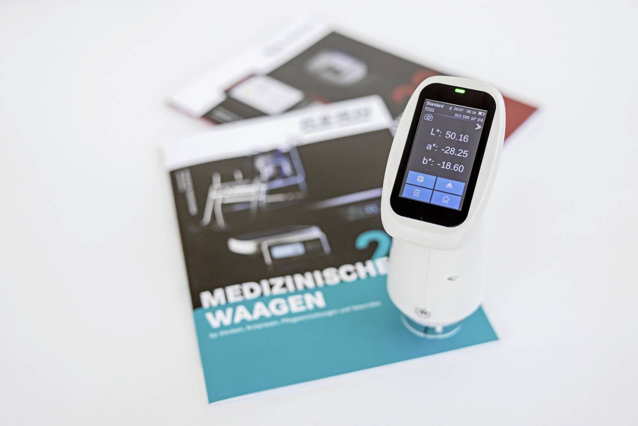 Ein medizinisches Gerät mit Display zeigt Messwerte an. Im Hintergrund liegen Broschüren mit dem Titel 'MEDIZINISCHE WAAGEN'.