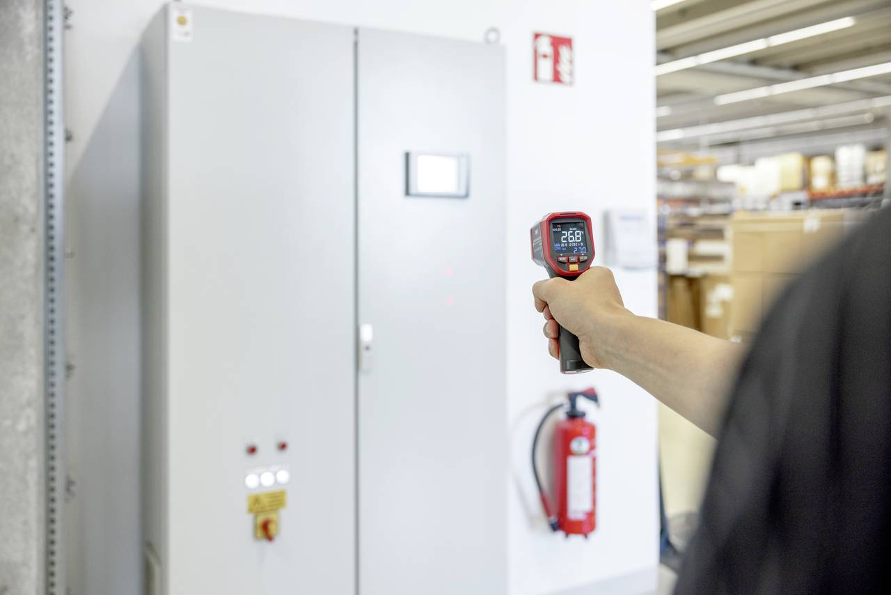 Eine Person misst die Temperatur eines grauen Schaltschrankes mit einem Infrarot-Thermometer, das 26,8 Grad Celsius anzeigt. Ein Feuerlöscher steht daneben.