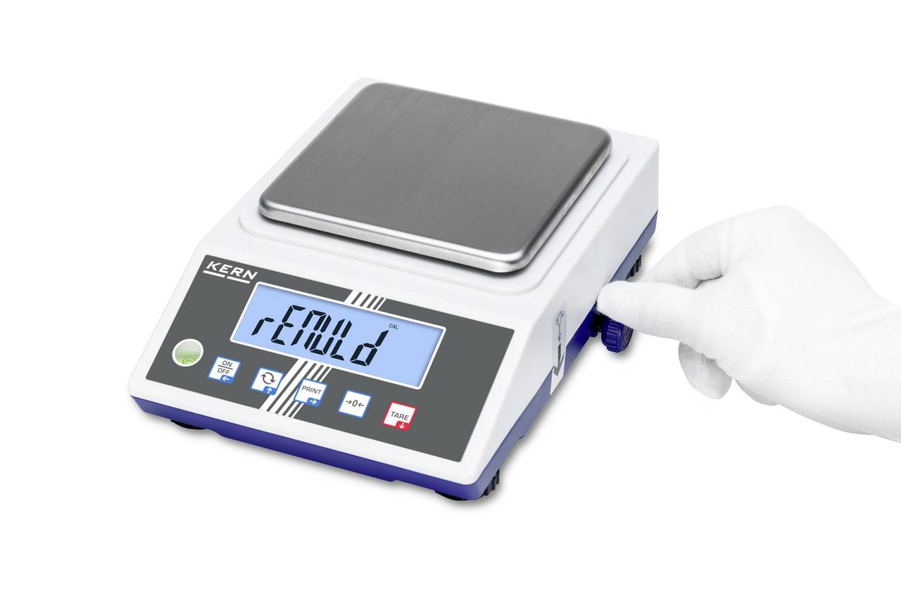 Digitale Laborwaage mit rechteckiger Edelstahlplattform und LCD-Anzeige. Eine handschuhtragende Hand bedient einen seitlichen Drehknopf.