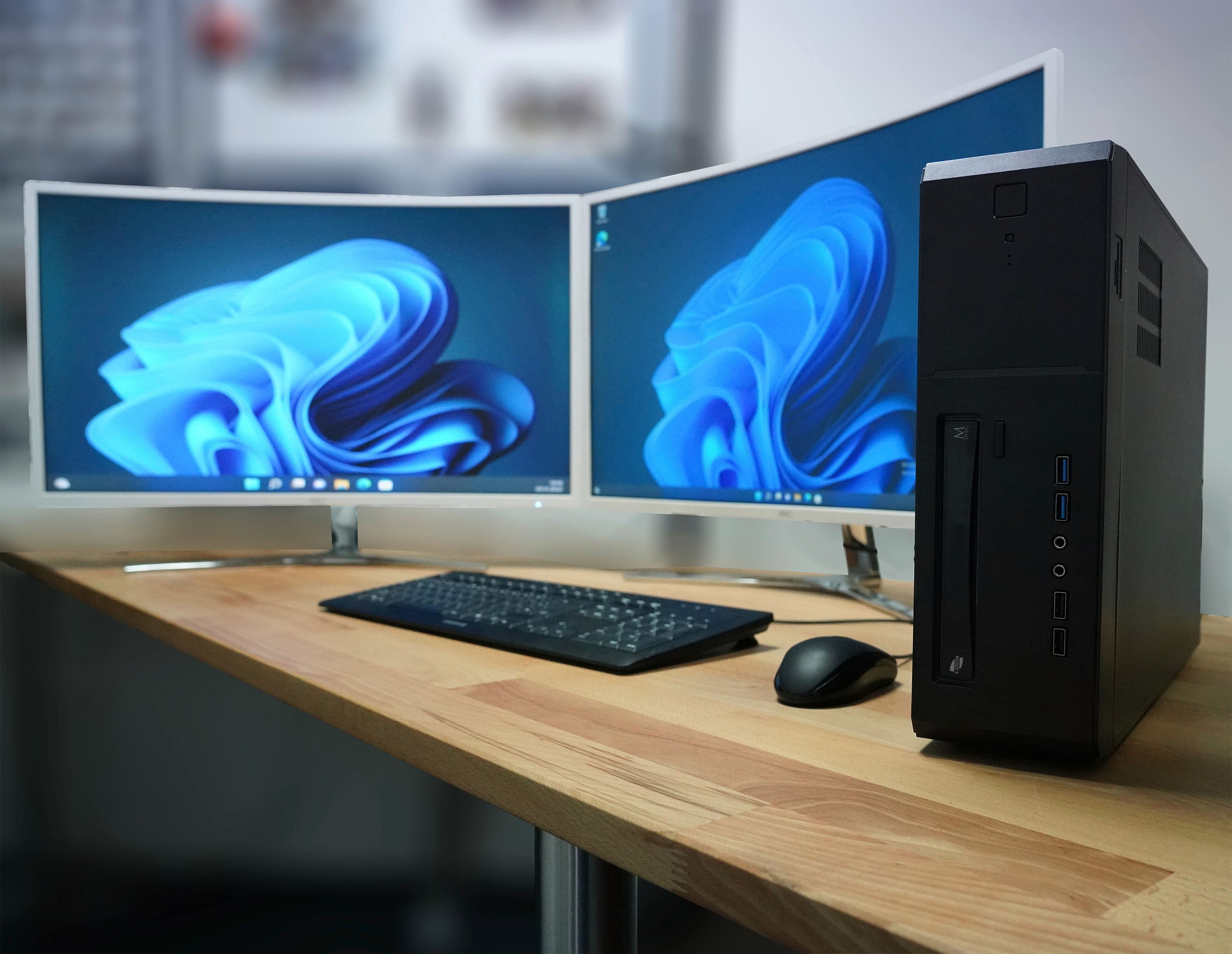 Ein Computerarbeitsplatz mit zwei gekrümmten Monitoren, einem schwarzen Computergehäuse, einer Tastatur und einer Maus auf einem Holztisch.