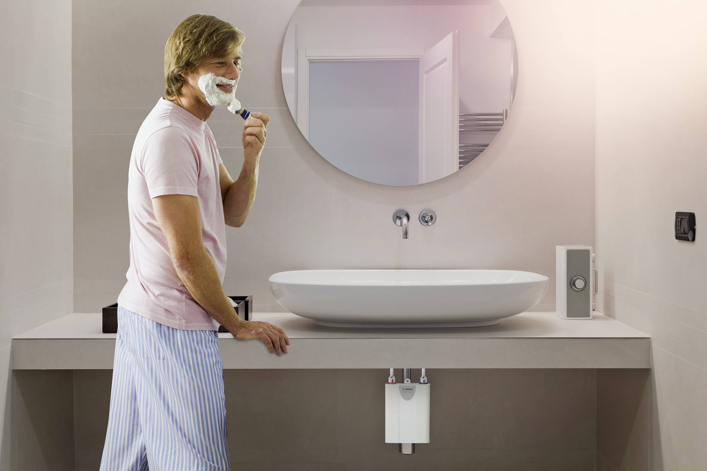 Ein Mann rasiert sich im Badezimmer vor einem Waschbecken und Spiegel. Er trägt ein rosa T-Shirt und gestreifte Schlafanzughosen.