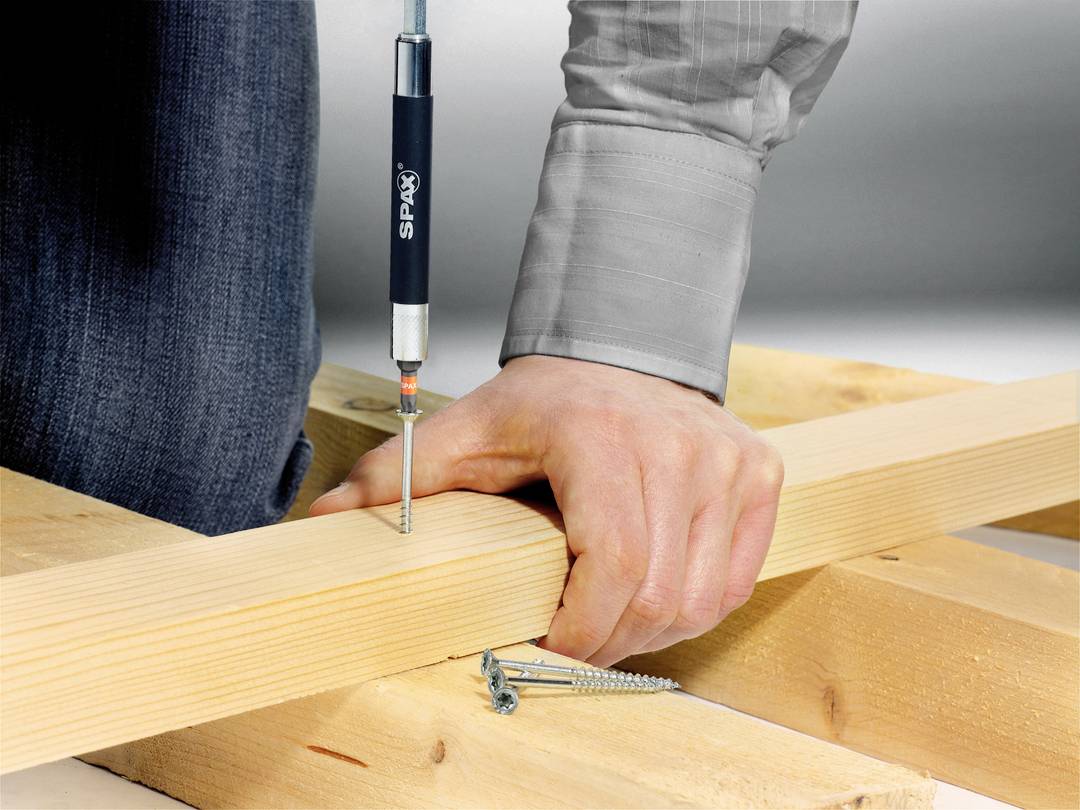 Eine Person schraubt mit einem Schraubendreher eine Schraube in ein Holzbrett. Mehrere Schrauben liegen daneben auf dem Holz.