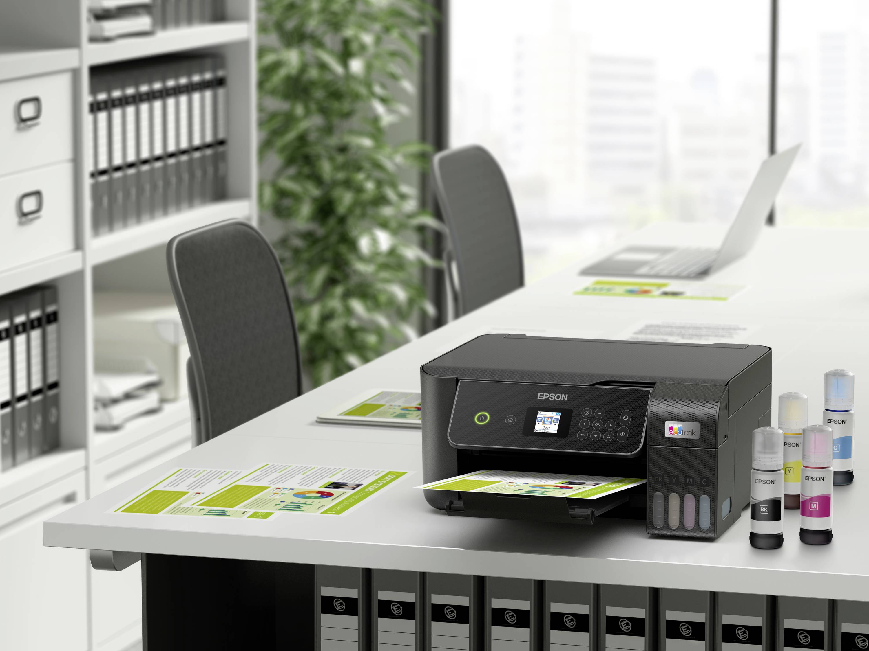 Ein Drucker auf einem Tisch in einem Büro druckt farbige Dokumente. Im Hintergrund sind Ordnerregale und ein Laptop zu sehen.