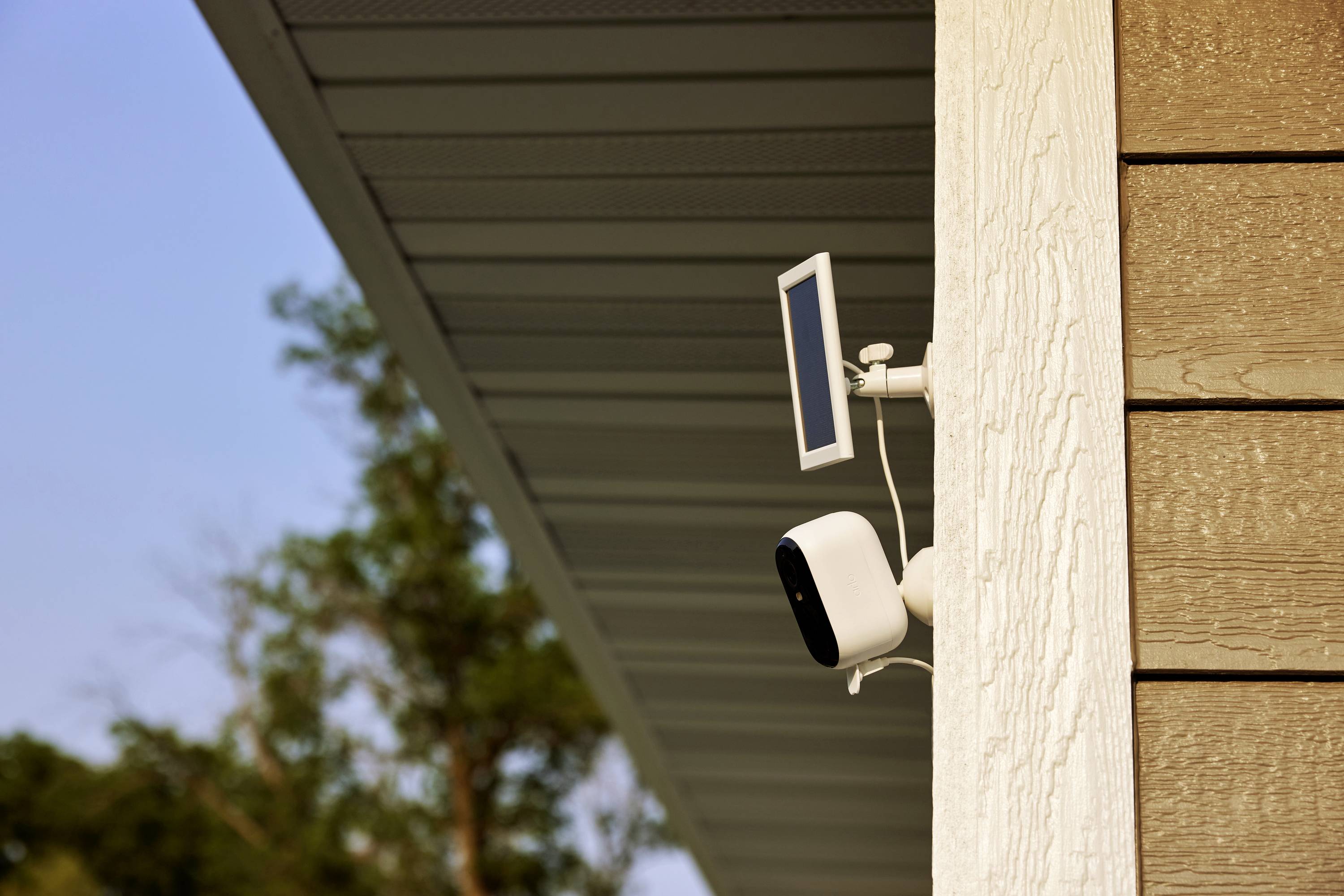 Eine Überwachungskamera ist an der Außenseite eines Gebäudes befestigt, mit einem kleinen Solarpanel darüber. Im Hintergrund sind Bäume sichtbar.