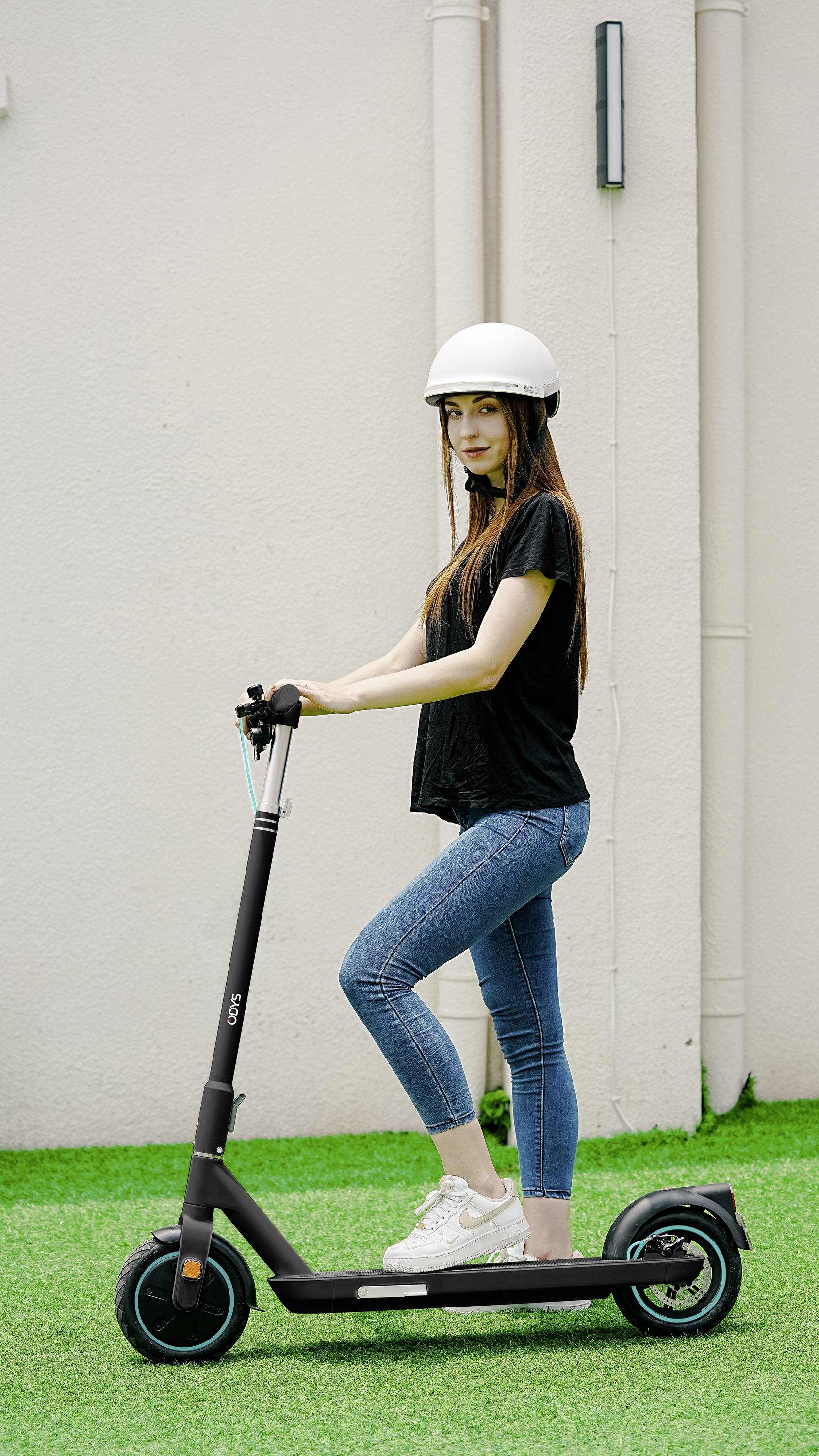 Eine Person steht mit einem weißen Helm auf einem elektrischen Roller, auf grünem Rasen vor einer hellen Wand.