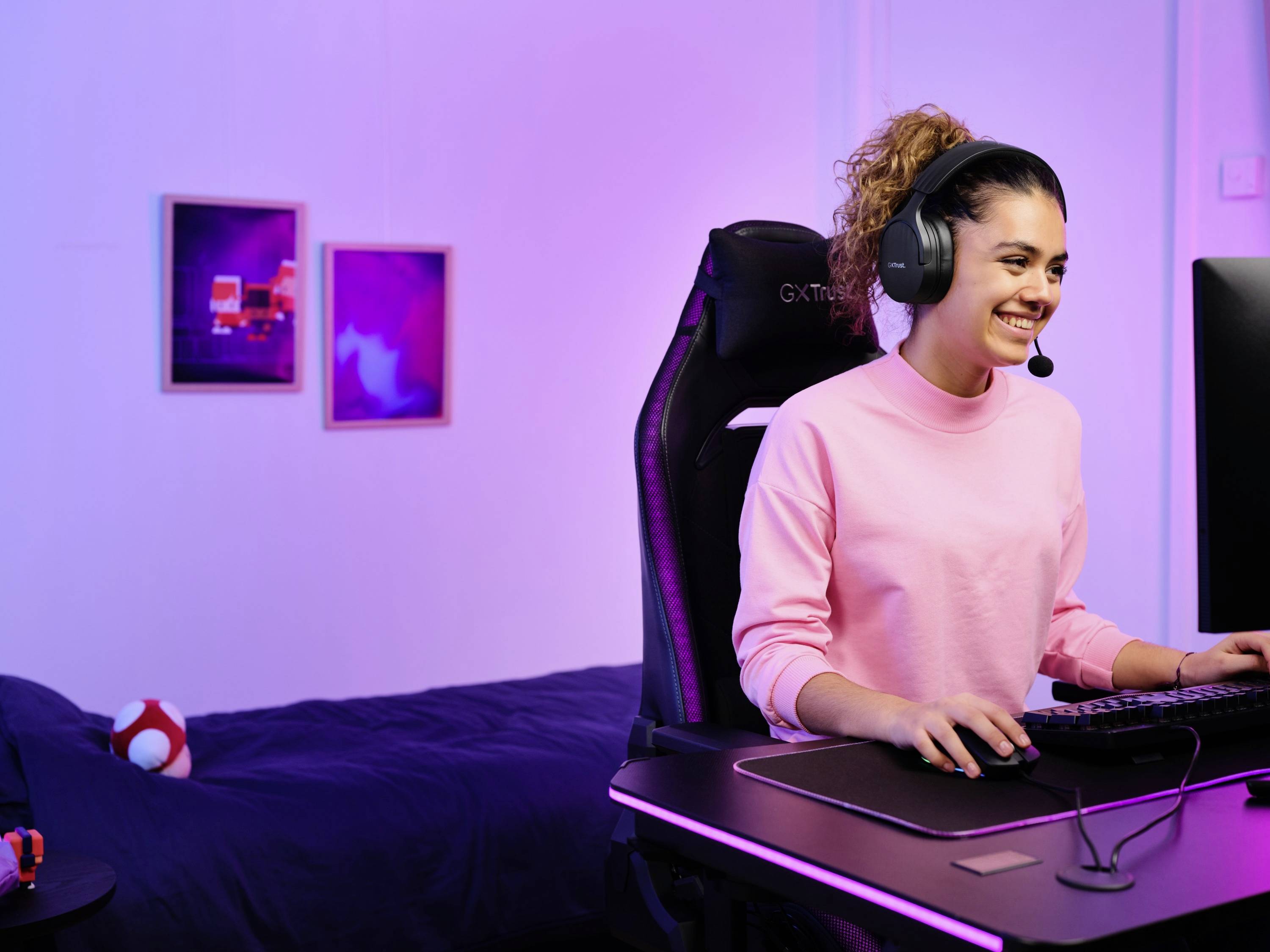 Eine Person mit Kopfhörern sitzt in einem Gaming-Stuhl vor einem Computer, lächelnd. Der Raum ist in violettes Licht getaucht.