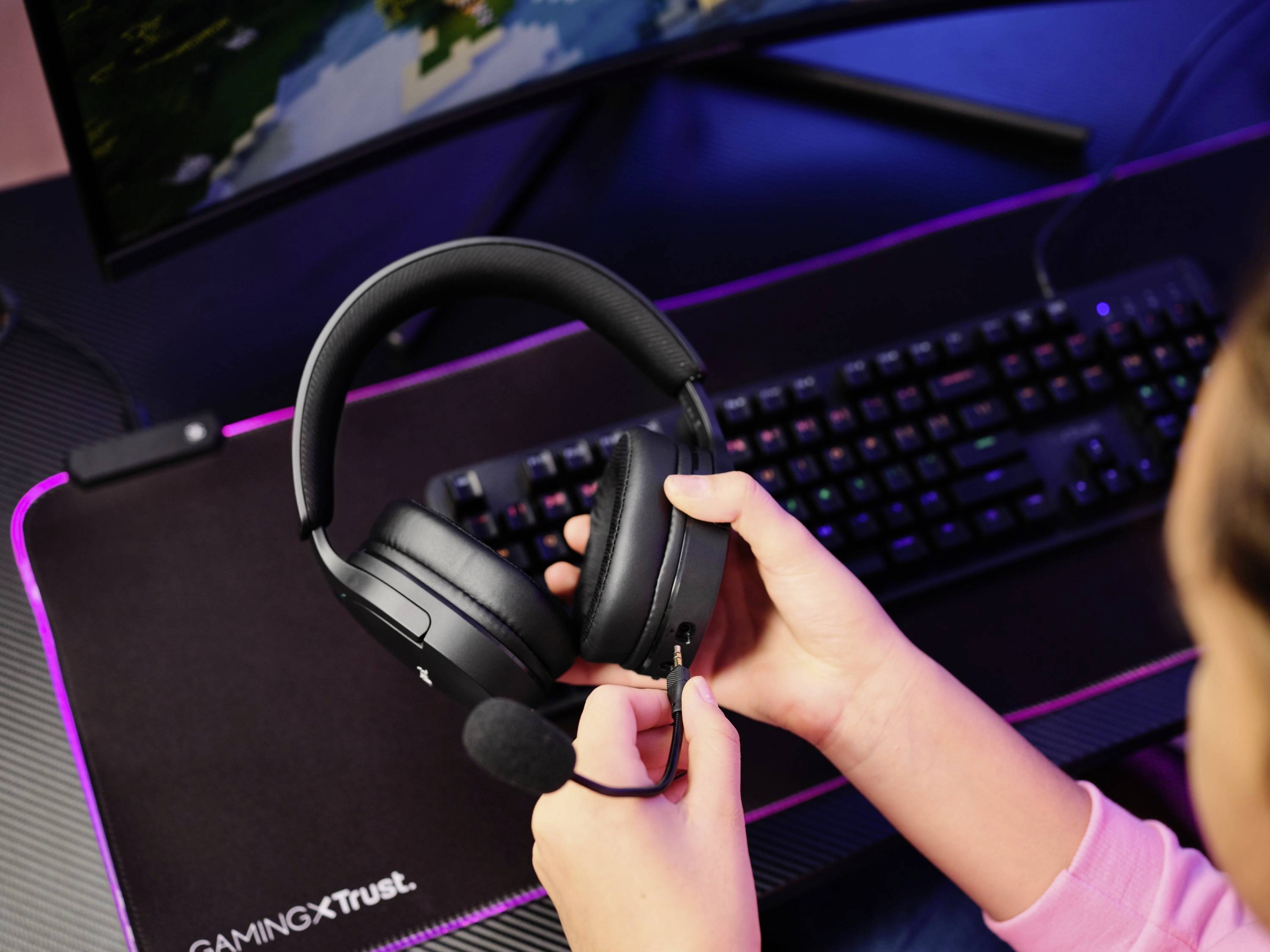 Eine Person hält ein Headset mit Mikrofon vor einem Computerbildschirm und einer beleuchteten Tastatur. Es scheint eine Gaming-Szene zu sein.