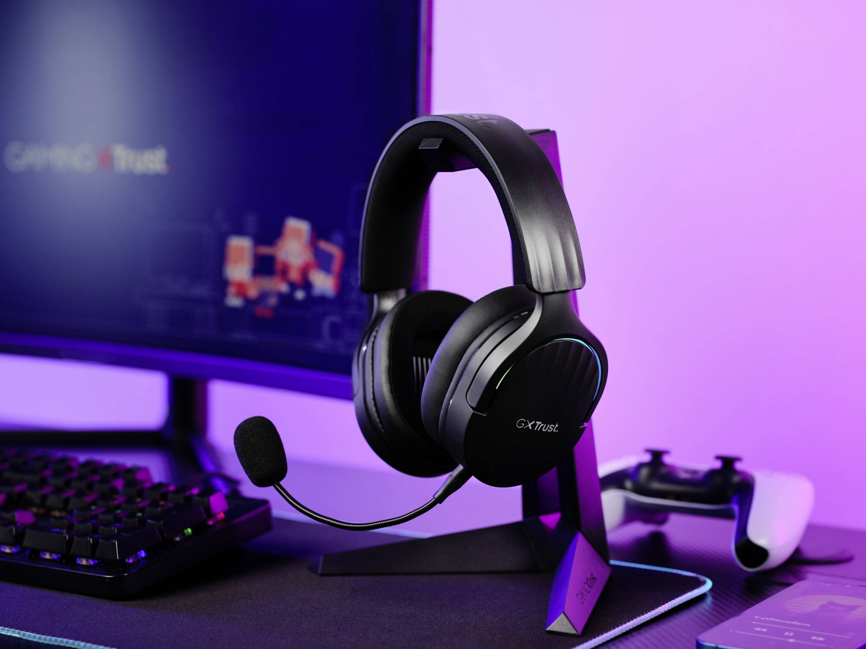 Ein Gaming-Headset mit Mikrofon steht auf einem Schreibtisch neben einem Monitor und einer Spielkonsole; alles in violetten Licht getaucht.