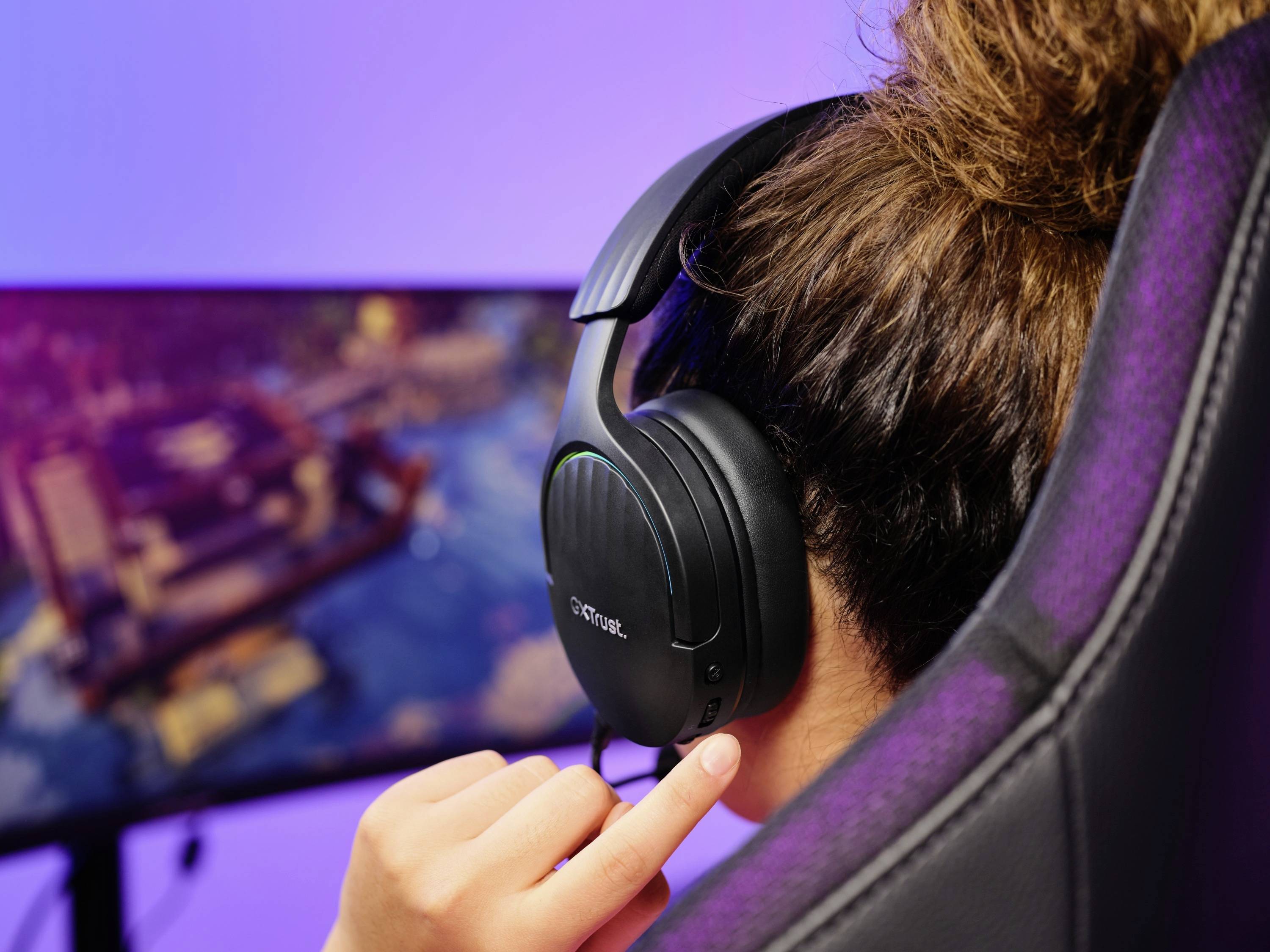 Eine Person sitzt vor einem großen Bildschirm und spielt ein Videospiel. Sie trägt ein Headset und hat eine Hand am Ohrhörer.