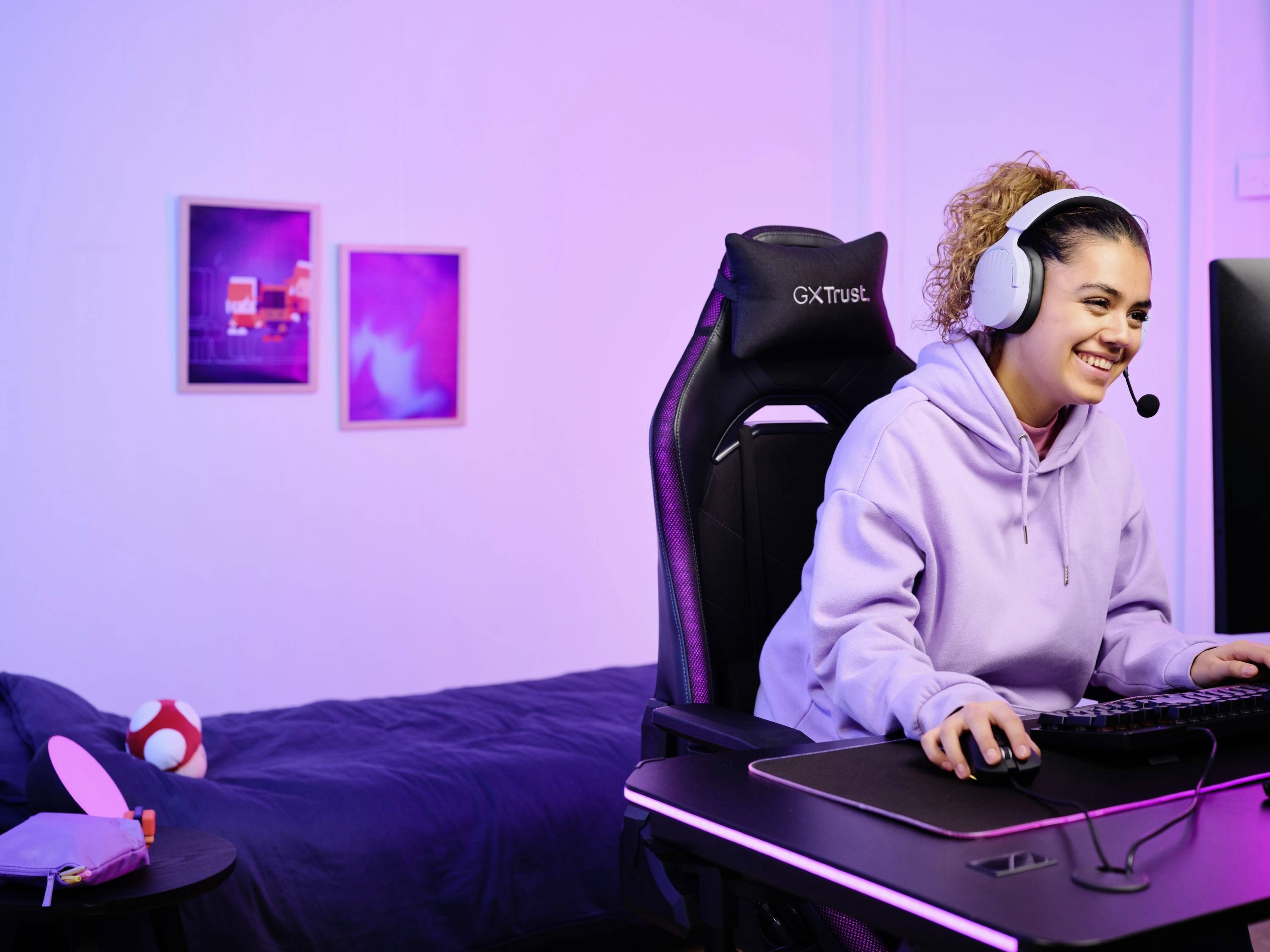 Eine Person mit Kopfhörern sitzt fröhlich an einem Computertisch in einem Schlafzimmer, konzentriert auf den Bildschirm.